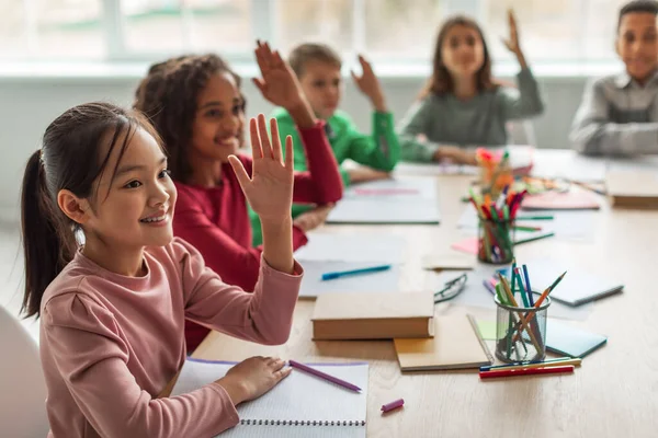 Happy Multiethnic School Kids Learning Raising Arms During Lesson Sitting At Desk In Classroom At School. Modern Education And Knowledge Concept. Selective Focus