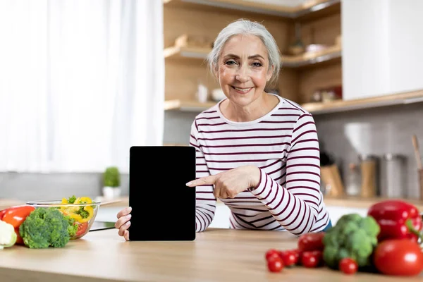 Smiling Elderly Lady Pointing At Blank Digital Tablet Screen In Kitchen, Mockup Image Of Happy Senior Woman Recommending New App Or Website While Cooking Healthy Food At Home, Copy Space