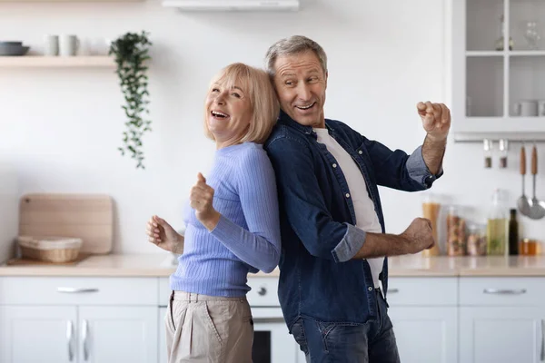 Joyful elderly spouses dancing together at kitchen, happy attractive senior man and woman in casual standing back to back and making dance moves, enjoying weekend at home, copy space