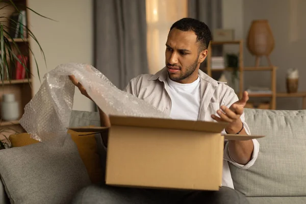 Dissatisfied black male customer opening box from online store, taking out packaging, unhappy with received item after unboxing parcel at home. Remote shopping, wrong delivery concept