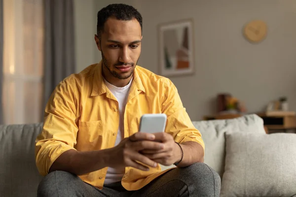 Serious millennial black man using cellphone, messaging online, sitting on sofa at home, copy space. Young African American guy with smartphone surfing internet. Gadget lifestyle, mobile apps