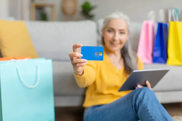 Happy caucasian retired gray-haired woman sits on floor with tablet and a lot of shopping bags, shows credit card in living room interior, blurred. Huge sale, online order, app, shopaholic at home