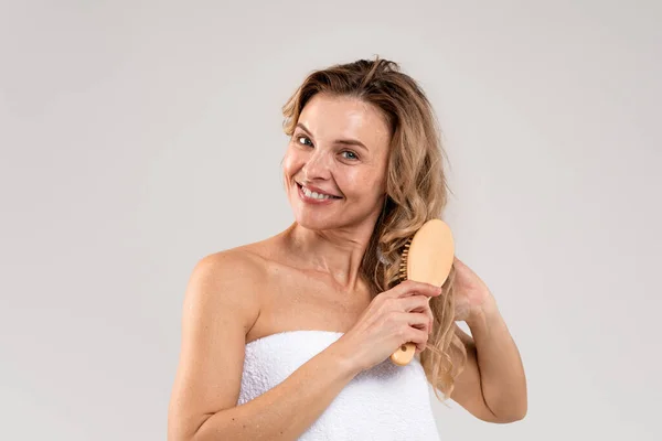 Beauty Routine. Attractive happy middle aged female brushing her blonde hair with bamboo comb, beautiful smiling woman standing wrapped in bath towel over light studio background, copy space