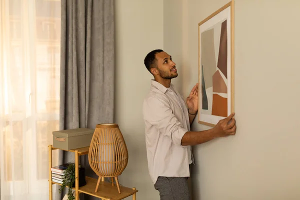 Young black man putting picture frame, hanging painting on wall, empty space. African American male interior designer decorating new modern stylish apartment. Home interior and domestic decor
