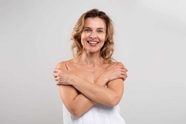 Enjoying Beauty. Happy Attractive Middle Aged Lady Wrapped In Bath Towel Embracing Herself, Beautiful Cheerful Female Smiling At Camera While Posing Over Light Grey Studio Background, Copy Space