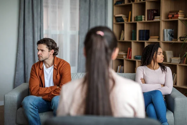 Upset angry young black wife ignores caucasian husband at meeting with doctor psychologist in office clinic interior. Family psychological therapy, treatment of mental problems, conflict and quarrel