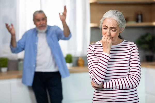 Psychological Abuse. Upset Senior Woman Crying In Kitchen While Arguing With Husband, Elderly Spouses Suffering Marital Crisis, Angry Man Standing On Background And Blaming Wife, Selective Focus