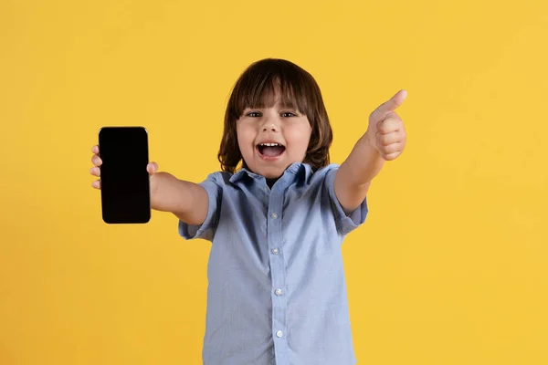Great kids content. Studio portrait of excited little boy showing smartphone with blank screen to camera and gesturing thumb up, yellow background, mockup
