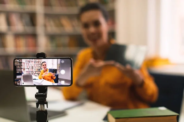 Female teacher shooting online lecture to students using cellphone on tripod and showing tablet, explaining something, selective focus on phone