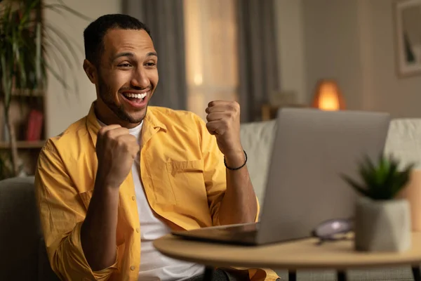 Excited young black man looking at laptop screen, gesturing YES, feeling happy about online win or business promotion at home. Millennial guy winning lottery or casino bet, gambling on pc