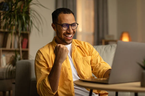 Joyful millennial black man with laptop celebrating online win or work success at home. Excited young African American guy enjoying big sale in web store, gambling on internet