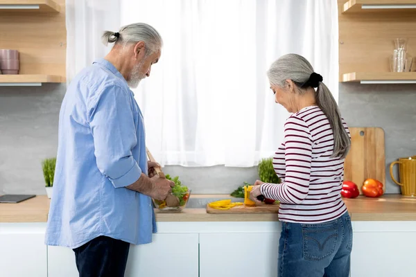 Cooking At Home. Portrait Of Senior Spouses Preparing Food Near Window In Kitchen, Back View Of Elderly Couple Making Vegetable Salad Together, Mature Man And Woman Enjoying Healthy Nutrition
