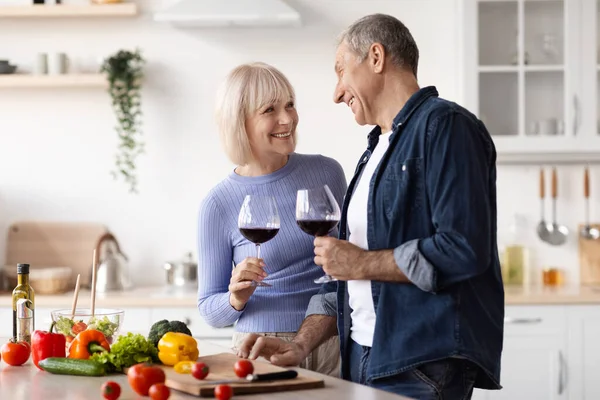 Positive elderly spouses drinking red wine while preparing meal at home, cutting fresh vegetables, holding glasses with drink, having conversation, enjoying time together, kitchen interior, copy space