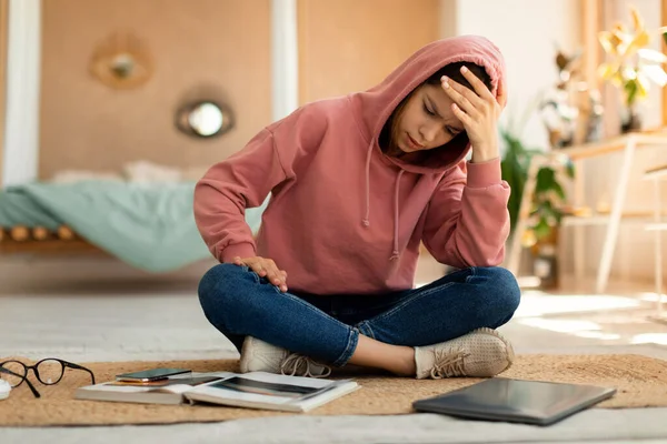 Exhautsed teenage girl doing homework alone, sitting on floor with laptop and book, touching her head, experiencing difficulties with studying