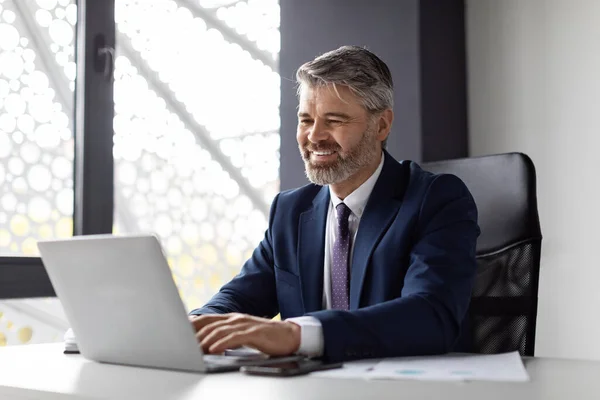 Portrait Of Smiling Middle Aged Businessman Working With Laptop Computer In Office, Handsome Male Entrepreneur In Suit Sitting At Desk And Typing On Keyboard, Enjoying Corporate Communication