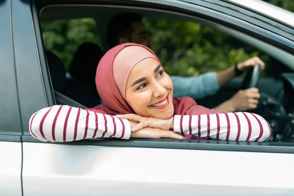 Happy glad young muslim man driving car with husband in hijab, lady looking out the window and enjoying car journey together, close up. New auto, safety in transport, life insurance and vacation trip