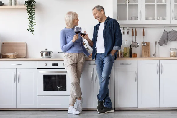 Happy senior couple drinking red wine at kitchen, elderly man and woman in casual outfits standing by kitchen table with glasses, having conversation, enjoying time together, copy space, full length