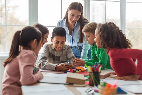 Multiethnic School Kids And Teacher Using Smartphone And Educational Application Learning Sitting At Desk In Modern Classroom At School. Education And Gadgets Concept. Selective Focus