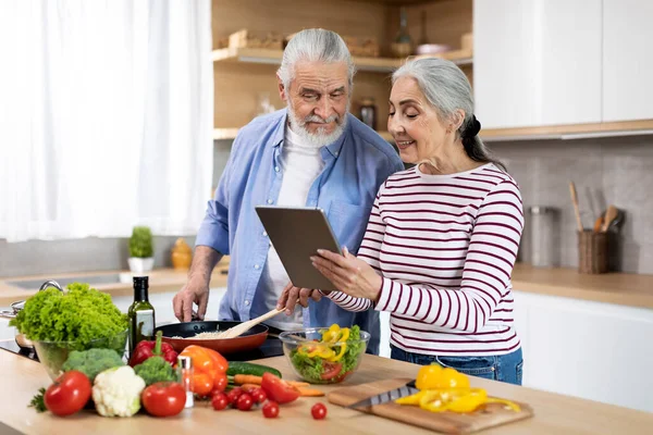 Happy Elderly Spouses Cooking Food In Kitchen And Using Digital Tablet Together, Married Senior Couple Checking Online Recipe While Preparing Healthy Vegetable Meal For Lunch At Home, Copy Space