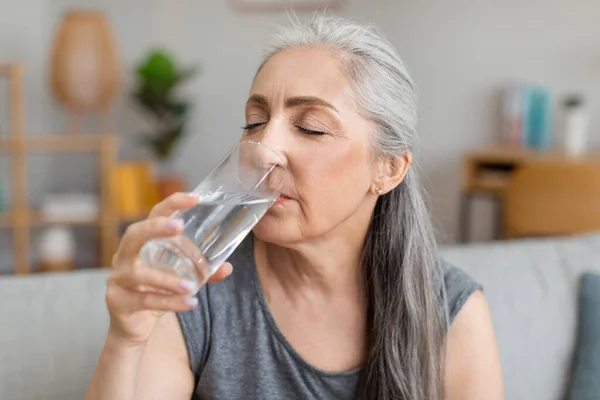 Caucasian elderly gray-haired woman drink water from glass, have useful habit in living room interior, close up. Aqua balance, thirst and prevent dehydration, healthy lifestyle, beauty care and diet