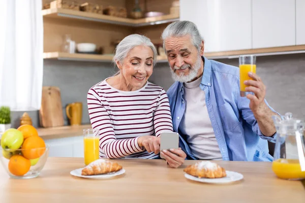 Happy Senior Couple Using Smartphone In Kitchen While Having Breakfast Together, Cheerful Elderly Spouses Shopping Online Or Browsing Social Networks On Cellphone And Enjoying Tasty Morning Meal