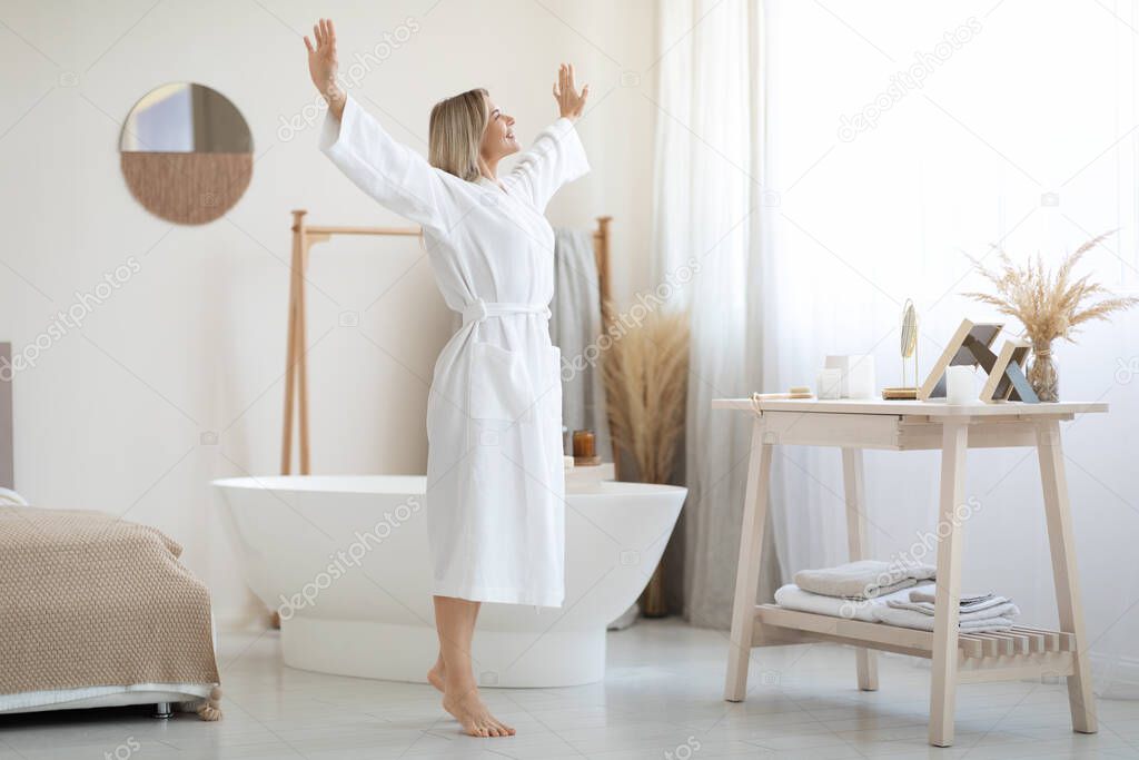 Full length shot of happy beautiful blonde woman in white bathrobe enjoying sunny morning, dancing by white modern open space bedroom, raising hands up and smiling, copy space
