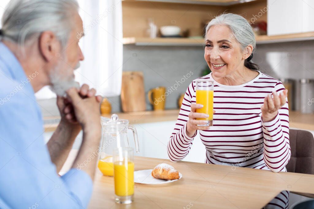 Happy Senior Spouses Chatting While Having Breakfast In Kitchen Together, Smiling Cheerful Elderly Couple Sitting At Table, Enjoying Homemade Pastry And Drinking Orange Juice, Closeup Shot