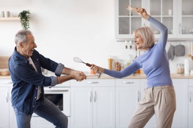 Happy senior couple enjoying time together at home, having fun at white cozy kitchen, cheerful elderly woman fighting with her husband as knights, using utensils as swords, copy space