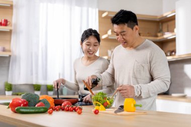Cheerful korean husband and wife making vegetable salad, adding olive oil, cooking healthy delicious dinner together at home, having conversation, cozy white kitchen interior, copy space