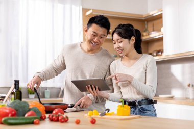 Food blog concept. Smiling asian couple checking recipes on Internet while making healthy dinner together at home, husband and wife looking at digital tablet screen, reading culinary blog, copy space