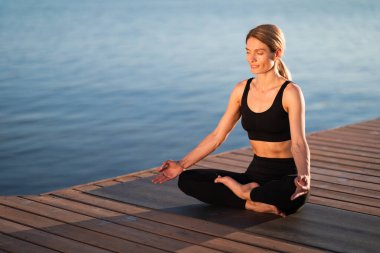 Wellbeing Concept. Beautiful Middle Aged Female In Activewear Meditating Outdoors, Portrait Of Calm Relaxed Woman Sitting In Lotus Position With Eyes Closed, Practicing Yoga On Wooden Pier Near River