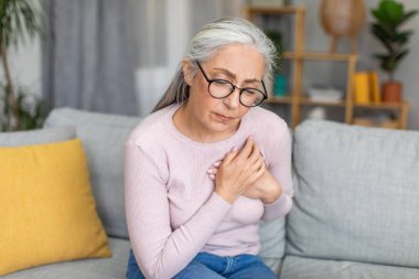 Sad caucasian elderly gray-haired woman in glasses presses her hands to chest and suffering from pain alone in living room interior. Health problems, heart attack, disease treatment and urgent help