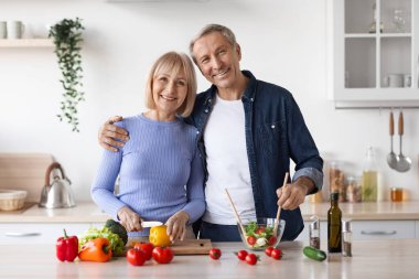 Attractive elderly man and woman in casual outfits cooking together at home, making delicious vegetable salad, embracing and smiling, kitchen interior, copy space. Healthy diet for seniors