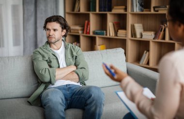 Dissatisfied young caucasian guy client ignores doctor at meeting with black lady psychologist in office clinic interior. Denial and psychological therapy, session with professional, help and support