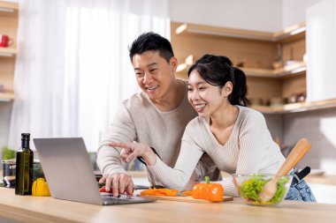 Positive japanese spouses watching culinary vlog while cooking healthy dinner, cheerful asian man and woman looking at laptop screen while preparing meal together at home, copy space