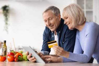 Senior couple using digital tablet and credit card, cooking together healthy vegetable salad at home, happy mature man and woman making lunch, order ingredients on Internet, copy space
