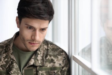 Postraumatic stress disorder in veterans concept. Depressed military young guy in camouflage uniform standing next to window, thinking of suicide, closeup photo, panorama with copy space