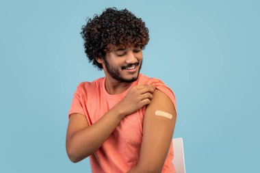 Happy curly young indian guy sitting on chair over blue studio background, looking at medical band on his shoulder and smiling, got vaccinated against flu or COVID-19, copy space, closeup