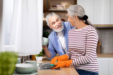Happy Senior Couple Spending Time Together In Kitchen At Home, Wife Washing Dishes While Husband Drinking Tea Next To Her, Cheerful Elderly Spouses Chatting And Making Domestic Chores, Closeup