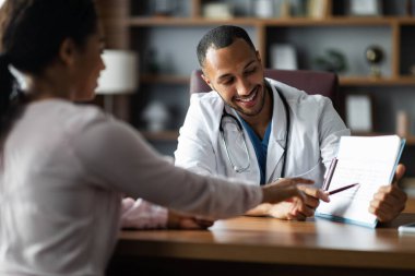 Cheerful handsome middle eastern young man doctor having conversation with black woman patient, practitioner showing treatment plan and smiling, giving details, modern clinic interior, copy space