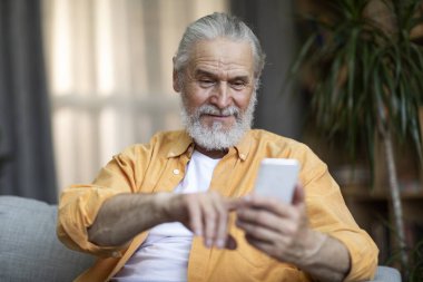 Lonely handsome bearded senior man sitting on couch at home, using brand new cell phone, touching gadget screen and smiling, using dating mobile application, copy space