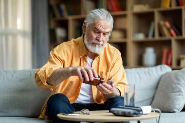 Sick senior man in casual outfit sitting on couch in living room, checking blood pressure and taking medicine, using tonometer, experiencing hypertension, home interior, copy space