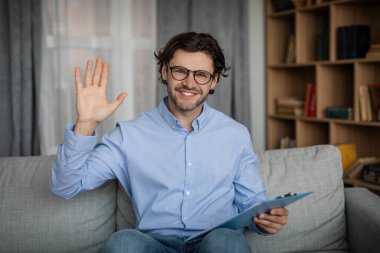 Glad cheerful young caucasian male doctor waving hand at camera in office interior. Online medical support, psychological therapy remotely, session with professional, video call and social distance