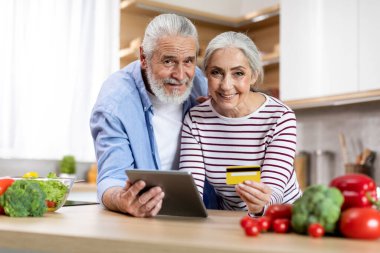 Portrait Of Smiling Elderly Spouses With Digital Tablet And Credit Card In Kitchen, Happy Married Senior Couple Using Modern Gadget For Online Shopping Or Payments In Internet, Copy Space