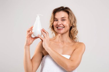 Beauty Ad. Smiling Middle Aged Woman Holding Bottle With Moisturising Body Lotion And Smiling At Camera, Beautiful Female Advertising New Cosmetic, Standing Wrapped In Towel On Grey Background