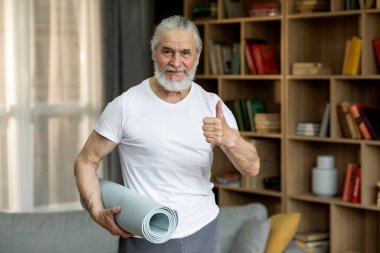 Cheerful grey-haired bearded senior man in sportswear exercising alone at home, holding yoga mat, showing thumb up and smiling at camera, copy space. Physical activity for seniors