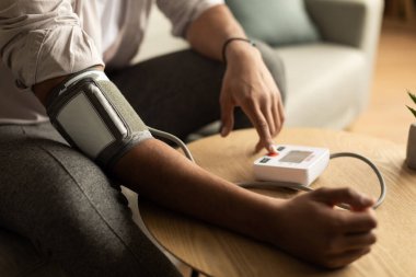 Closeup of young black man checking blood pressure, using modern tonometer at home. Unrecognizable African American male having hypertension, feeling unwell, cropped view