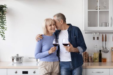 Romantic senior couple drinking red wine at kitchen, happy elderly man and woman in casual outfits standing by kitchen table with glasses, hugging and cuddling, enjoying time together, copy space