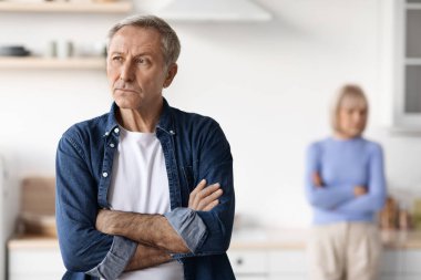 Selective focus on upset mature man with arms crossed on chest looking aside, having quarrel with his wife, kitchen interior. Crisis in marriage for seniors, divorce at older ages concept