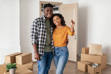 Black Husband And Wife Holding New Home Key Moving To House Hugging Standing Among Cardboard Boxes Indoor, Smiling To Camera. Real Estate Buyers Concept. Selective Focus